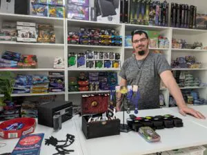 Der Inhaber des Geschäfts steht hinter seiner Ladentheke und präsentiert eine umfangreiche Auswahl an Sammelkarten und Figuren.